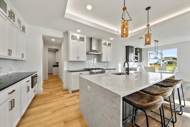 kitchen featuring wall chimney exhaust hood, sink, a raised ceiling, a kitchen island with sink, and white cabinets