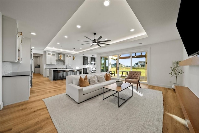 living room with ceiling fan, a raised ceiling, and light wood-type flooring