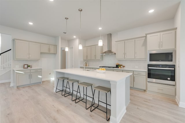 kitchen featuring gray cabinetry, a kitchen island with sink, wall chimney range hood, light hardwood / wood-style flooring, and appliances with stainless steel finishes