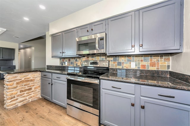 kitchen with gray cabinetry, light hardwood / wood-style flooring, backsplash, dark stone countertops, and appliances with stainless steel finishes