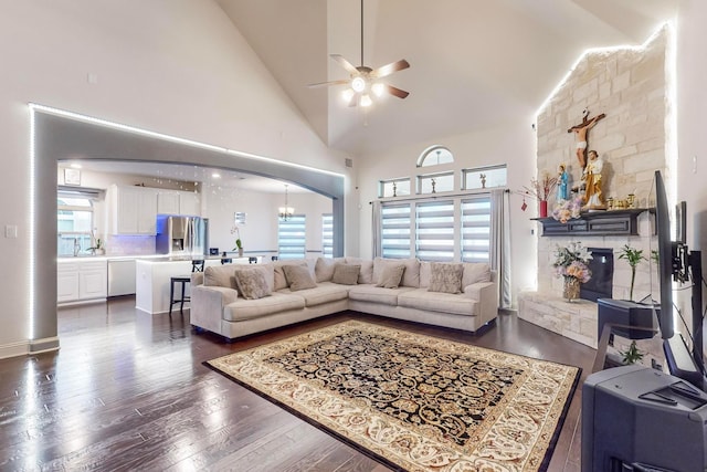 living room featuring a stone fireplace, dark wood-type flooring, ceiling fan with notable chandelier, and high vaulted ceiling