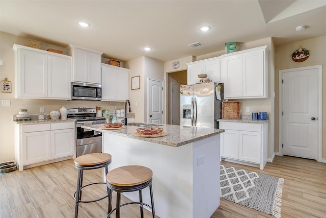 kitchen with appliances with stainless steel finishes, white cabinetry, light stone countertops, a center island with sink, and light wood-type flooring