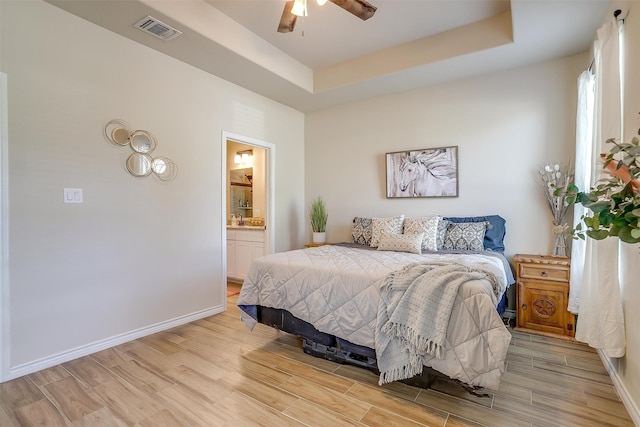 bedroom featuring connected bathroom, ceiling fan, light hardwood / wood-style floors, and a tray ceiling