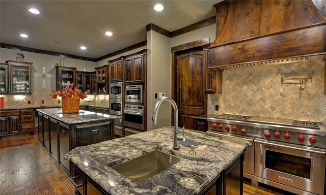 kitchen with sink, a kitchen island with sink, dark brown cabinetry, custom exhaust hood, and range with two ovens