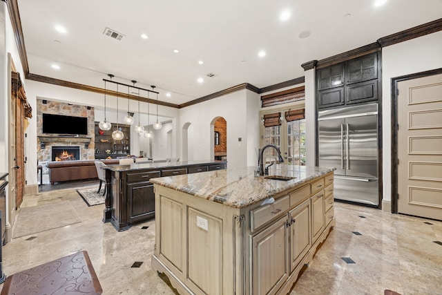 kitchen featuring stainless steel built in refrigerator, a kitchen island with sink, sink, cream cabinets, and a stone fireplace