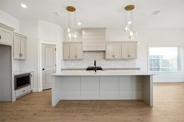 kitchen with a center island with sink, decorative light fixtures, stainless steel microwave, and light wood-type flooring