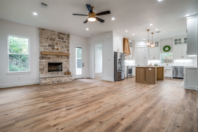 unfurnished living room featuring ceiling fan with notable chandelier, light hardwood / wood-style floors, a stone fireplace, and a wealth of natural light