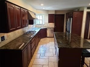 kitchen with stainless steel appliances, range hood, backsplash, dark stone countertops, and a kitchen bar