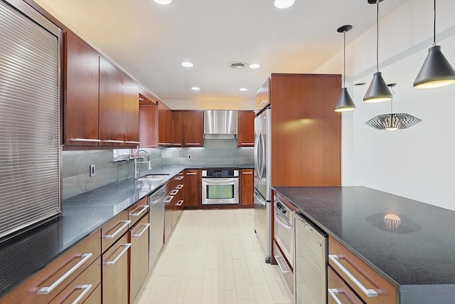 kitchen with sink, appliances with stainless steel finishes, dark stone countertops, hanging light fixtures, and wall chimney exhaust hood