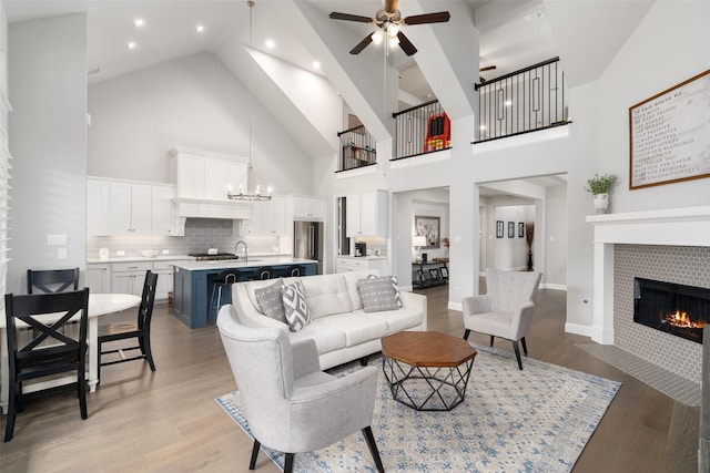 living room with sink, a tile fireplace, high vaulted ceiling, ceiling fan with notable chandelier, and light wood-type flooring