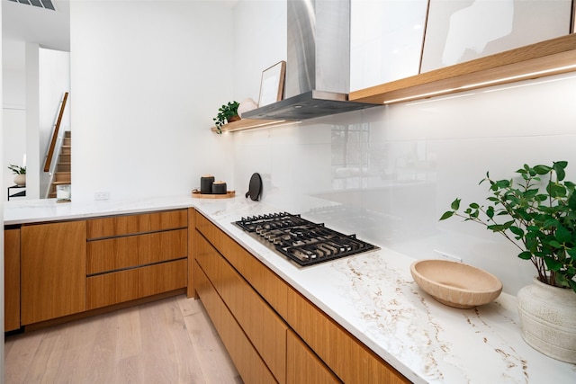 kitchen with stainless steel gas cooktop, decorative backsplash, light wood-style floors, wall chimney range hood, and light stone countertops