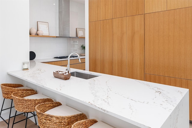 kitchen featuring light stone counters, open shelves, a breakfast bar, a sink, and wall chimney exhaust hood