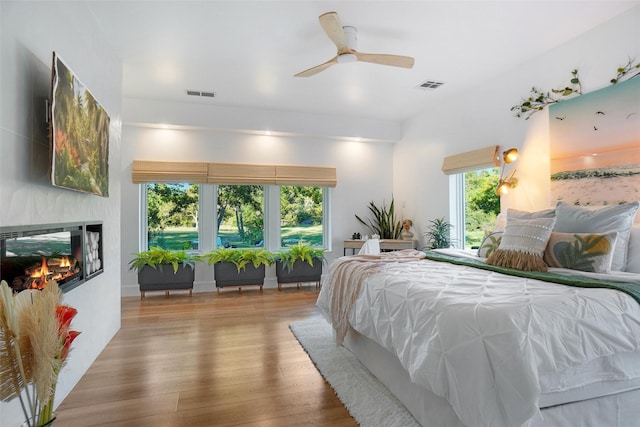 bedroom with wood finished floors, a glass covered fireplace, visible vents, and a ceiling fan