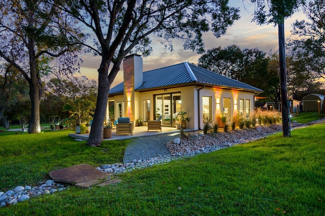 back of property at dusk with metal roof, a lawn, stucco siding, a chimney, and a patio area