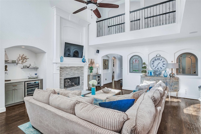 living room with ceiling fan, dark wood-type flooring, a fireplace, a high ceiling, and wine cooler