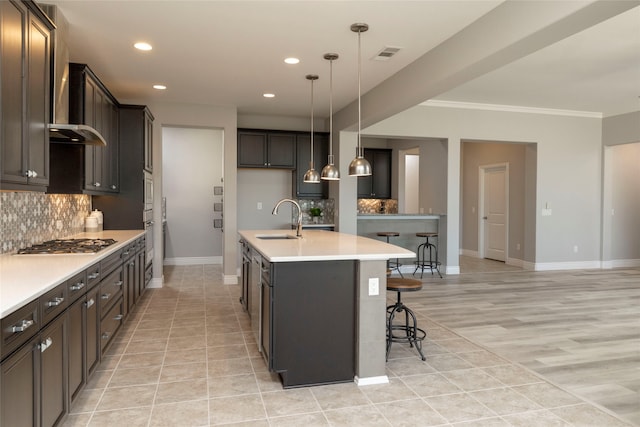 kitchen with light wood-type flooring, ornamental molding, sink, decorative light fixtures, and an island with sink