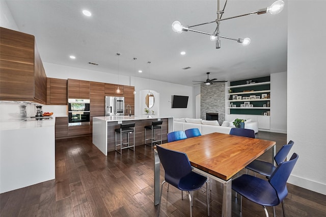 dining space with a stone fireplace, sink, dark hardwood / wood-style flooring, built in features, and ceiling fan with notable chandelier