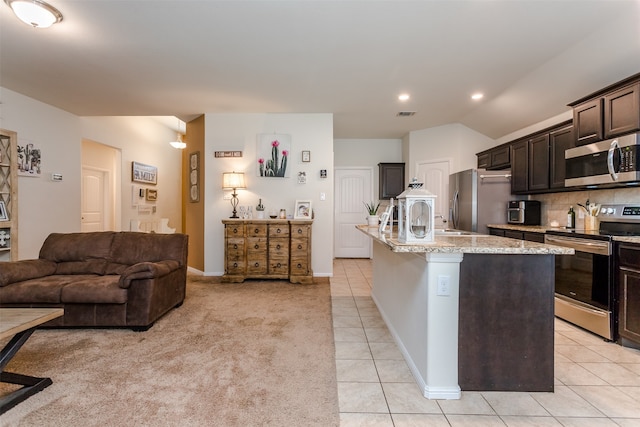 kitchen with light stone countertops, an island with sink, light carpet, dark brown cabinets, and appliances with stainless steel finishes