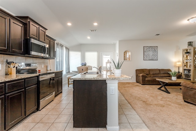 kitchen featuring light stone counters, an island with sink, light colored carpet, dark brown cabinets, and appliances with stainless steel finishes