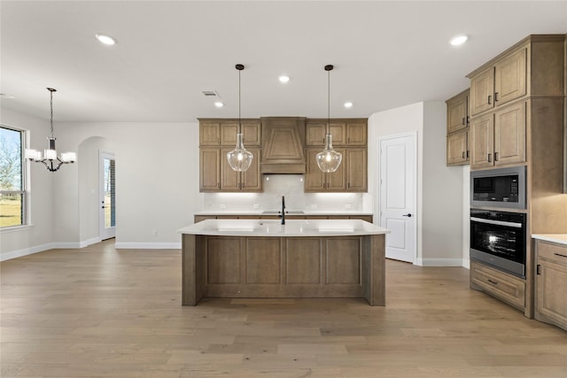 kitchen featuring a center island with sink, premium range hood, oven, built in microwave, and light wood-type flooring