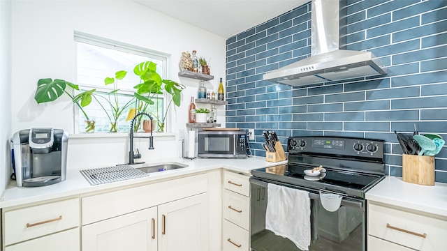 kitchen featuring black electric range oven, sink, white cabinetry, wall chimney range hood, and backsplash