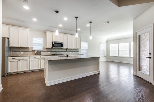 kitchen featuring appliances with stainless steel finishes, dark wood-type flooring, decorative light fixtures, white cabinetry, and an island with sink