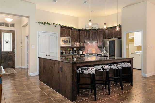 kitchen with washer / clothes dryer, appliances with stainless steel finishes, dark brown cabinets, and a kitchen island with sink