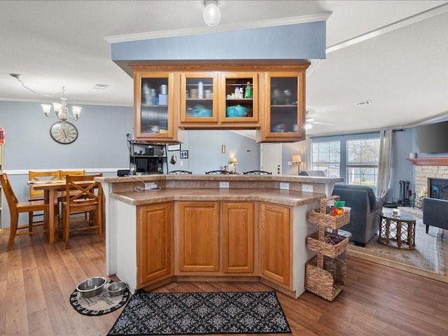 kitchen featuring crown molding, ceiling fan with notable chandelier, light hardwood / wood-style floors, a stone fireplace, and kitchen peninsula