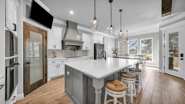 kitchen featuring light stone countertops, a kitchen island with sink, white cabinets, custom range hood, and appliances with stainless steel finishes