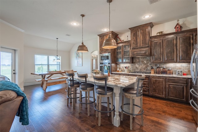 kitchen featuring appliances with stainless steel finishes, dark brown cabinetry, tasteful backsplash, an island with sink, and decorative light fixtures