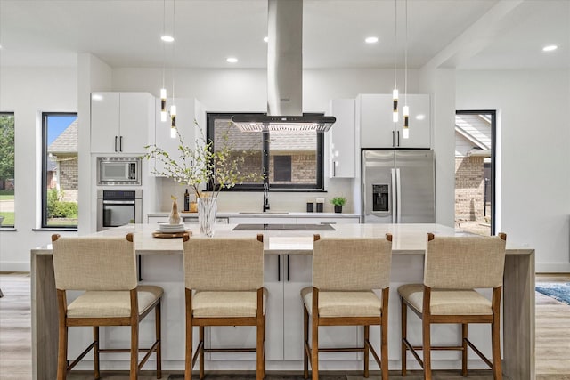 kitchen with hanging light fixtures, stainless steel appliances, island range hood, white cabinets, and a kitchen bar