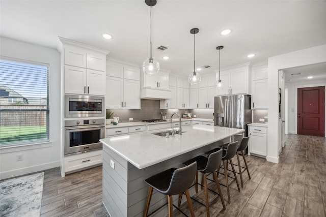 kitchen featuring appliances with stainless steel finishes, wood-type flooring, a center island with sink, decorative light fixtures, and white cabinets