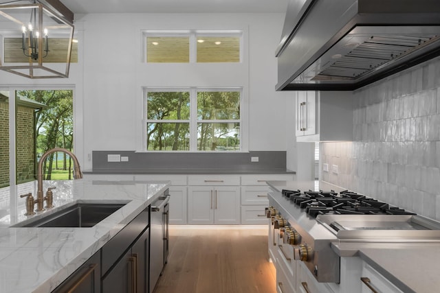 kitchen featuring a wealth of natural light, sink, wall chimney range hood, light stone counters, and white cabinets