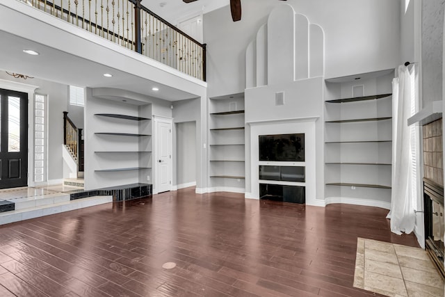 unfurnished living room featuring a high ceiling, built in features, ceiling fan, and a tiled fireplace