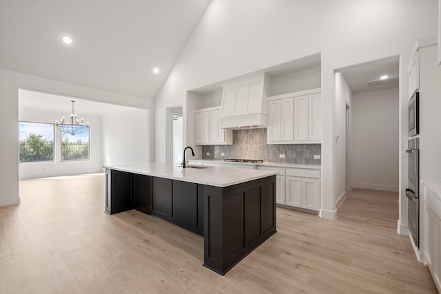 kitchen featuring premium range hood, an island with sink, sink, white cabinets, and hanging light fixtures