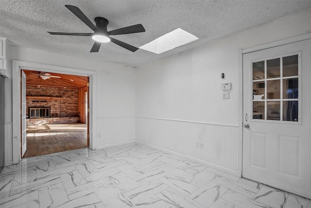 unfurnished room featuring ceiling fan, a brick fireplace, a textured ceiling, and a skylight