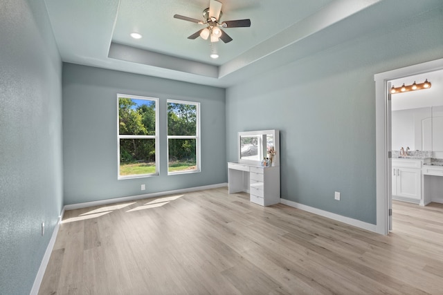 unfurnished bedroom featuring light hardwood / wood-style flooring, ensuite bath, ceiling fan, and a tray ceiling