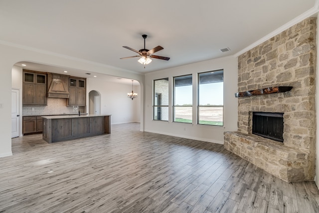 unfurnished living room with ceiling fan with notable chandelier, light hardwood / wood-style flooring, a stone fireplace, and ornamental molding