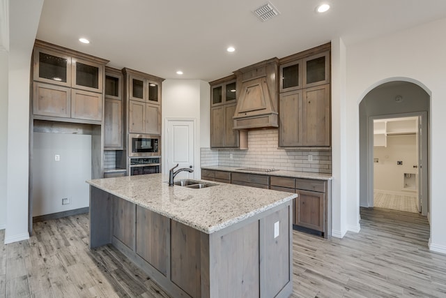 kitchen with built in microwave, sink, stainless steel oven, light stone counters, and a kitchen island with sink