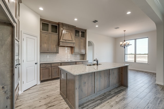 kitchen featuring light stone counters, custom range hood, sink, decorative light fixtures, and a center island with sink