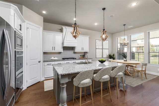 kitchen featuring white cabinetry, light stone counters, appliances with stainless steel finishes, pendant lighting, and a kitchen island with sink