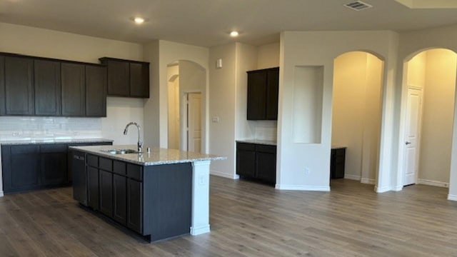 kitchen featuring sink, hardwood / wood-style flooring, light stone countertops, a center island with sink, and decorative backsplash