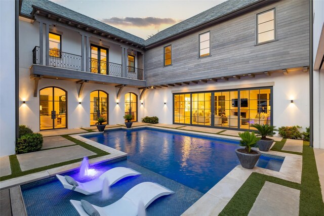 back house at dusk with a patio area, a balcony, a swimming pool with hot tub, and french doors