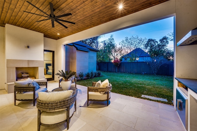 patio terrace at dusk with ceiling fan, a yard, and an outdoor fireplace