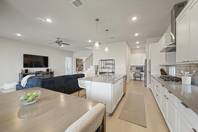 kitchen with an island with sink, sink, white cabinets, stainless steel appliances, and wall chimney range hood