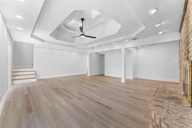 unfurnished living room with a brick fireplace, a textured ceiling, a tray ceiling, ceiling fan, and light hardwood / wood-style flooring