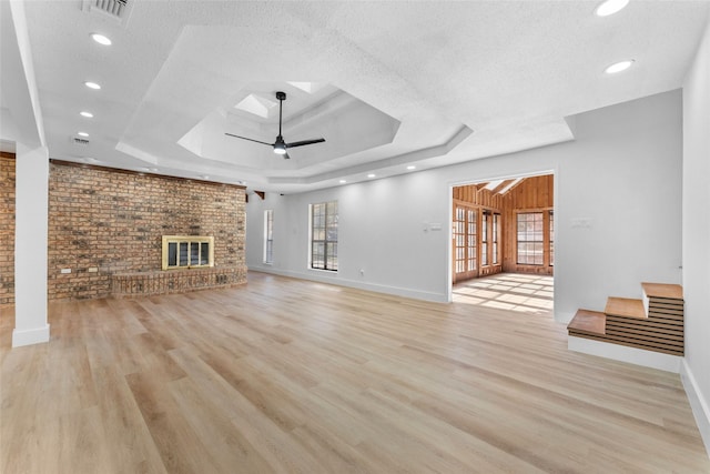 unfurnished living room featuring ceiling fan, a brick fireplace, brick wall, light hardwood / wood-style floors, and a tray ceiling