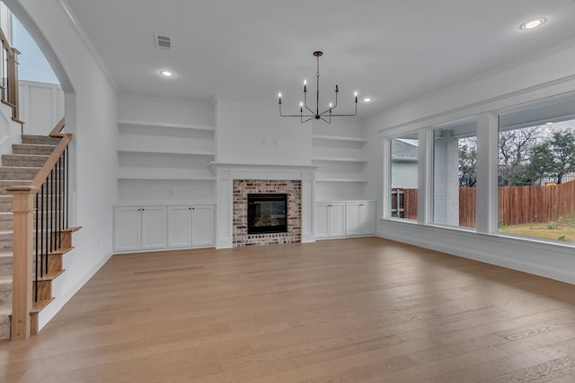 unfurnished living room featuring a brick fireplace, crown molding, built in features, an inviting chandelier, and light hardwood / wood-style flooring