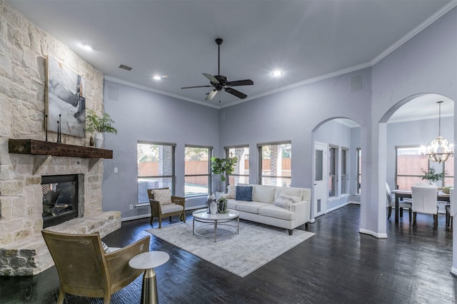 living room with dark wood-type flooring, a stone fireplace, crown molding, a towering ceiling, and ceiling fan with notable chandelier