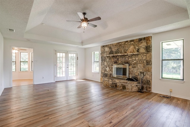 unfurnished living room featuring light wood-type flooring, a textured ceiling, a raised ceiling, ceiling fan, and a stone fireplace